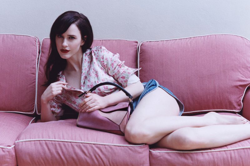 Brunette woman in denim shorts and a floral shirt poses with a handbag on a pink sofa in an indoor setting. Fashion photography.
