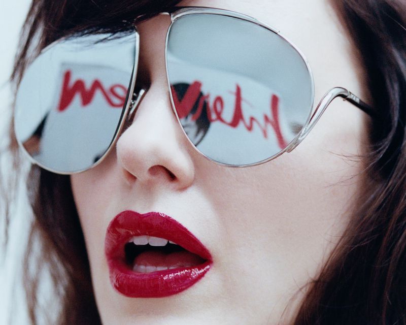 Close-up of a woman with glossy red lips wearing silver sunglasses reflecting red text against a white background.