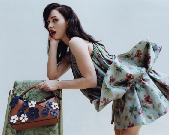 Attractive brunette poses with a brown handbag adorned with white and blue flowers, wearing a floral dress, leaning on a green chair.