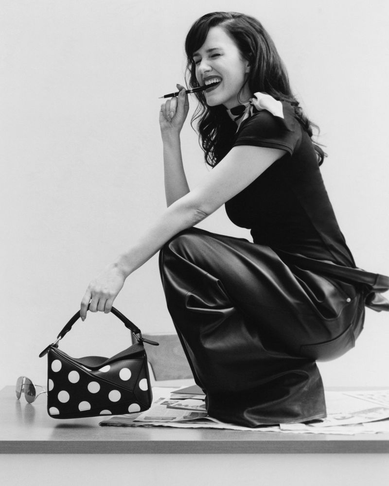 A joyful woman with dark hair holds a polka-dot purse and a pen in a black-and-white studio portrait, smiling and crouching.