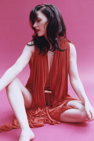 Rachel Brosnahan sitting barefoot in a draped rust-orange plunging gown against a vivid pink studio backdrop.