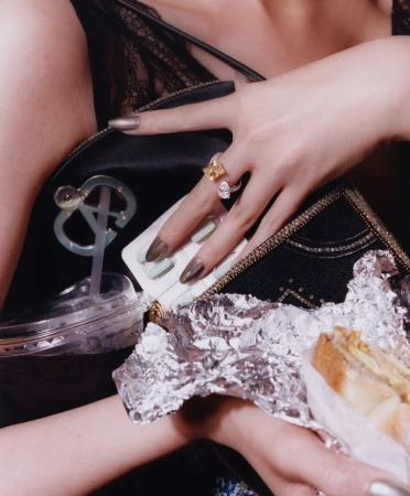 Close-up of manicured hands holding a black handbag, a drink cup, and foil-wrapped food for WSJ Girl Grip Goes Glam.