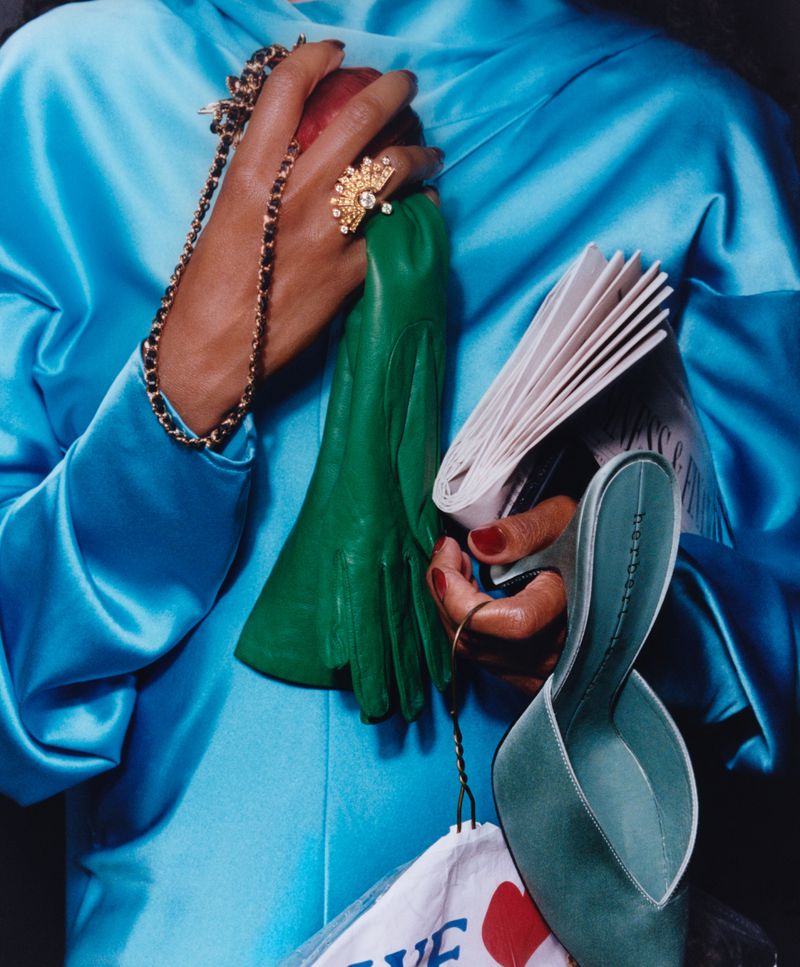 Close-up of hands holding a green leather glove, gold ring, chain-strap handbag, and a turquoise mule shoe against a bright blue top.
