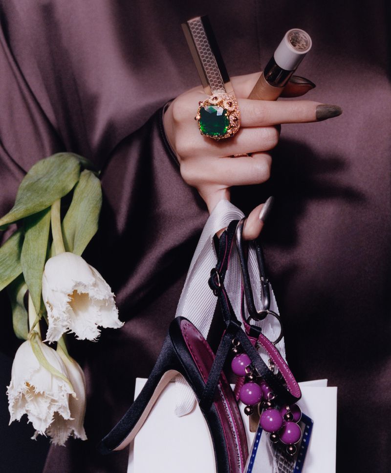 Close-up of a person holding two lipsticks, a large green ring, white tulips, a shopping bag, and black stiletto heels against a satin garment.