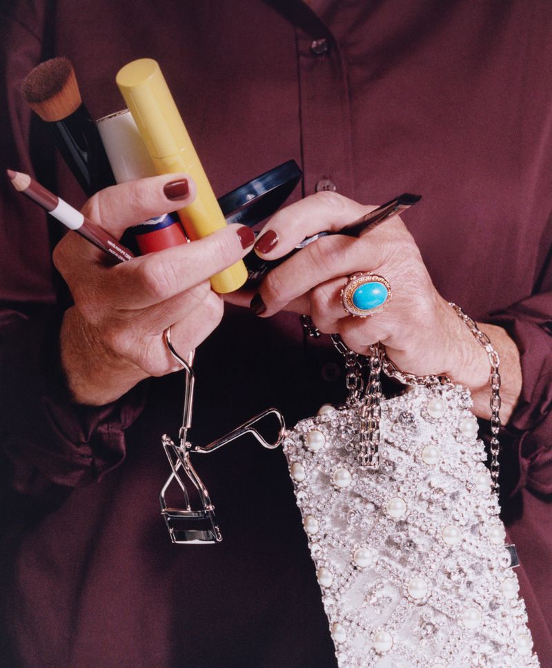 Close-up of hands holding makeup brushes, a yellow tube, and a silver jewelled handbag, wearing a brown shirt and a turquoise ring.
