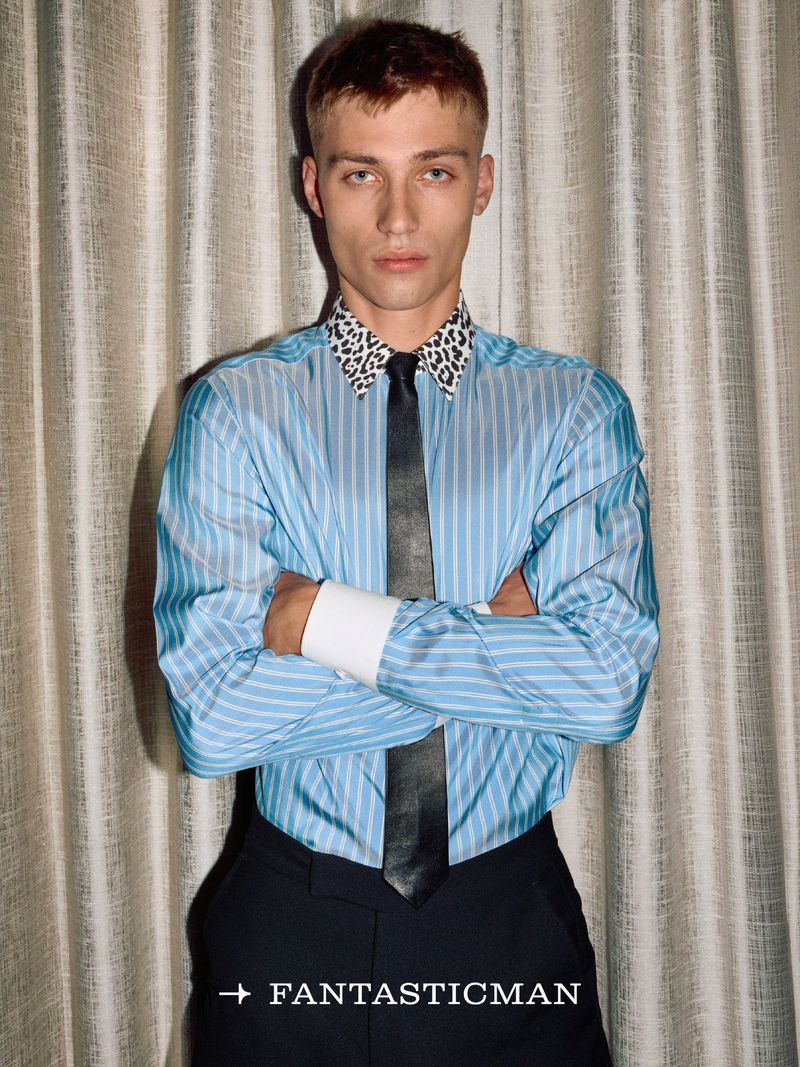 Young man with short brown hair wearing a blue striped shirt with a black tie against a textured curtain background.