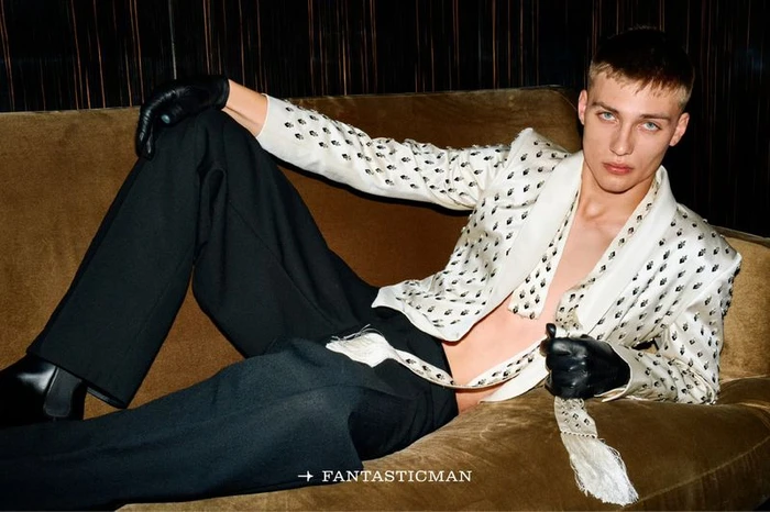 A young male model reclining on a brown sofa wearing a white patterned jacket, black gloves, and dark trousers.
