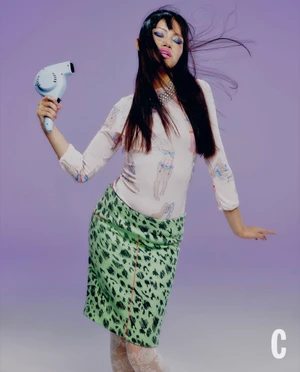 Model holding a blue hairdryer in front of a purple gradient background wearing a patterned top and green skirt