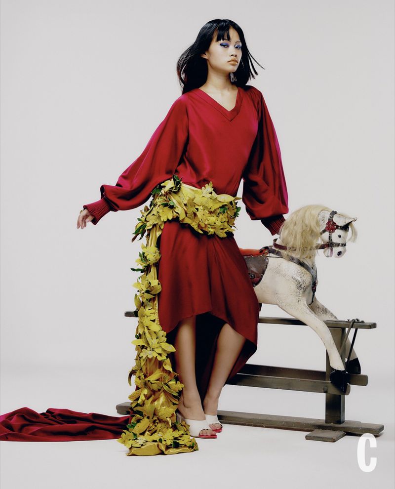 Model in red dress with floral sash sitting on a rocking horse against a white backdrop
