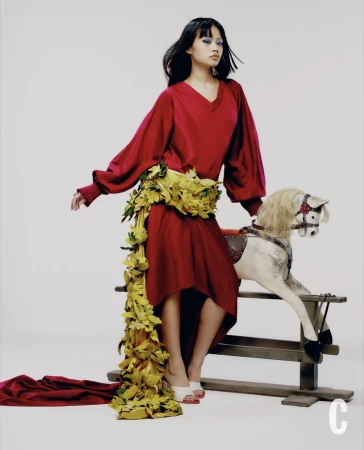 Model in red dress with floral sash sitting on a rocking horse against a white backdrop