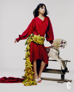 Model in red dress with floral sash sitting on a rocking horse against a white backdrop