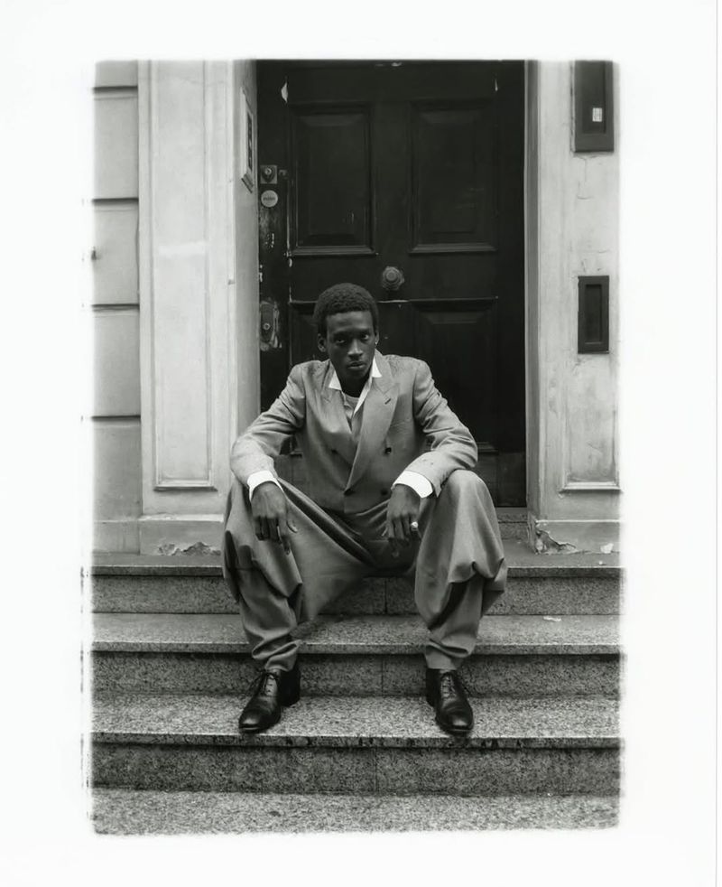 A black man in a double-breasted suit seated on stone steps before a dark wooden door for Financial Times HTSI Magazine.