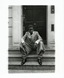 A black man in a double-breasted suit seated on stone steps before a dark wooden door for Financial Times HTSI Magazine.