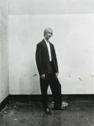 Monochrome portrait of a male model wearing a dark blazer, white shirt, and trousers against a textured wall.