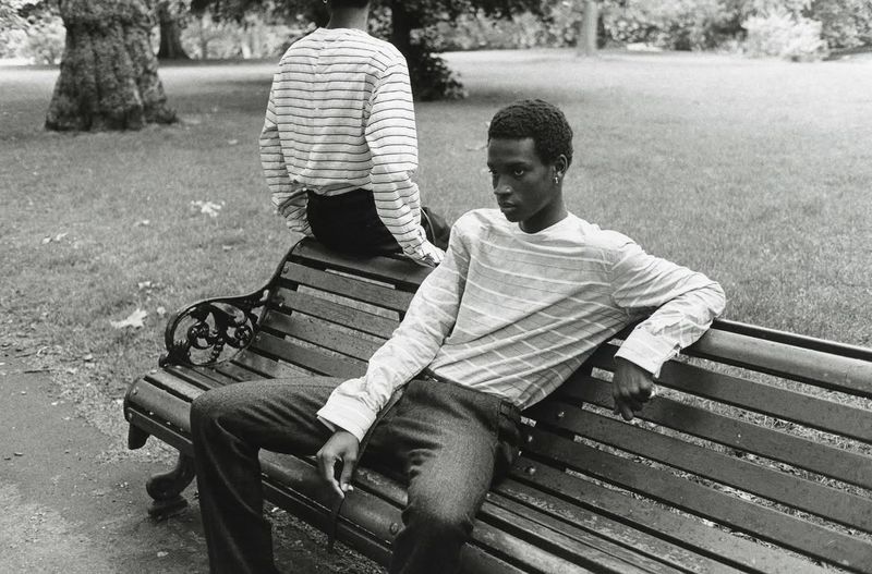 Two men seated on a park bench wearing striped long-sleeve shirts and trousers in a black and white editorial fashion photograph.