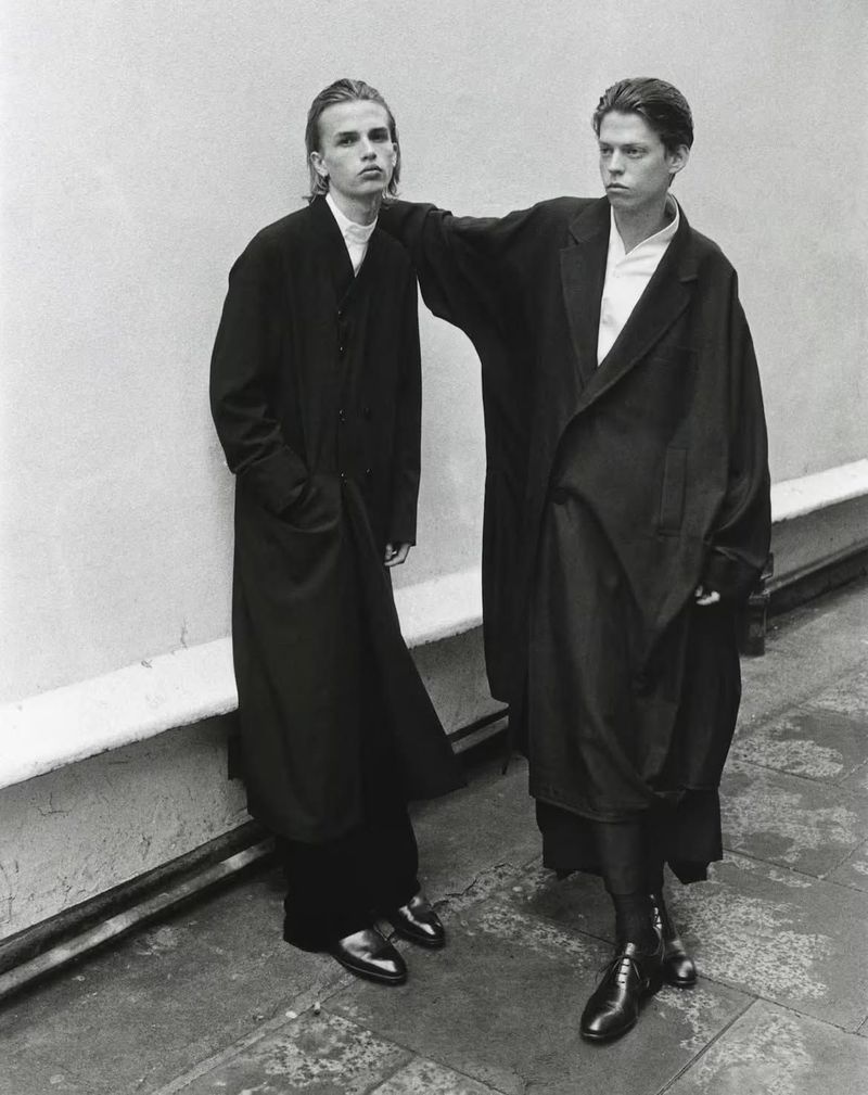Two young male models wearing long black coats standing against a light wall for an editorial by Kingsley Ifill.