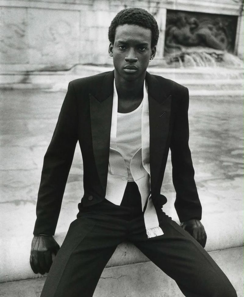 Black man wearing a black tuxedo jacket and vest with a white shirt, seated outdoors near a stone structure.