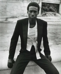 Black man wearing a black tuxedo jacket and vest with a white shirt, seated outdoors near a stone structure.