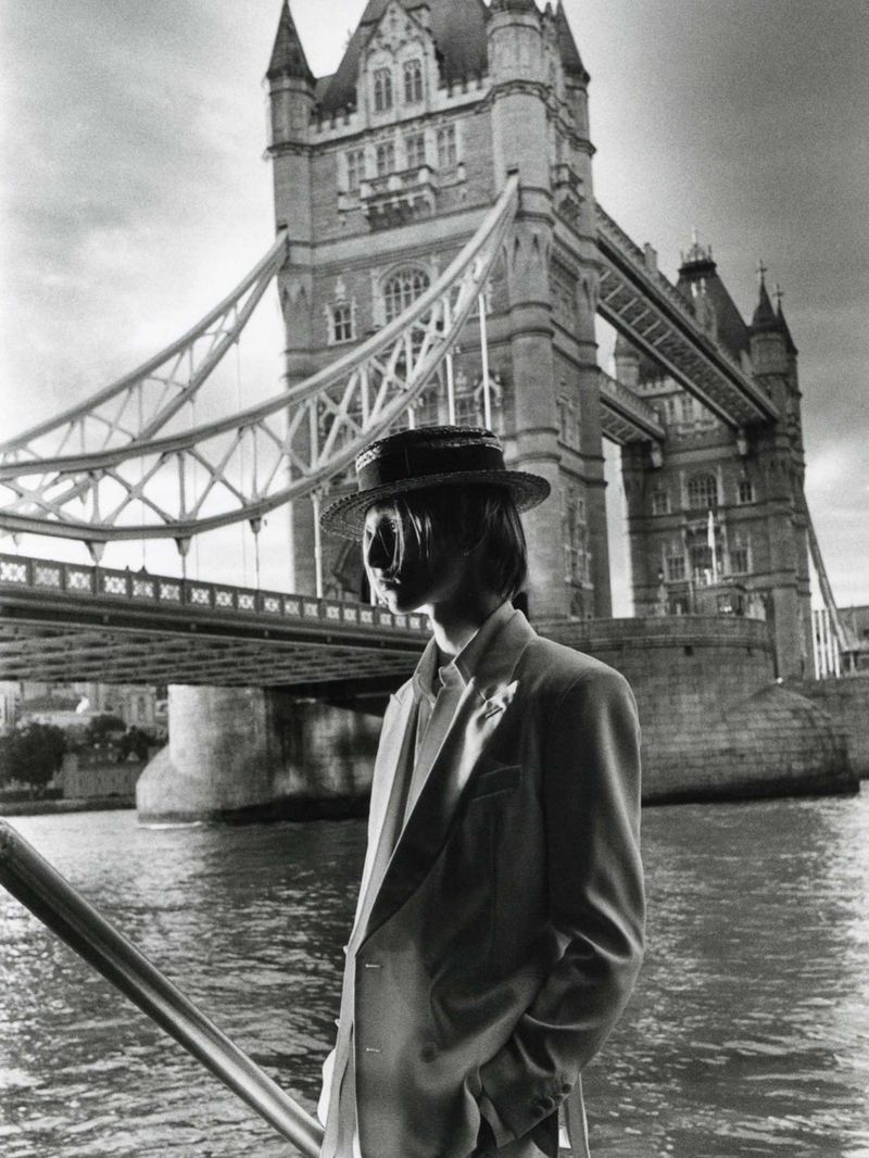 Monochrome portrait of a person in a tailored suit and straw hat standing by water with Tower Bridge in the background.