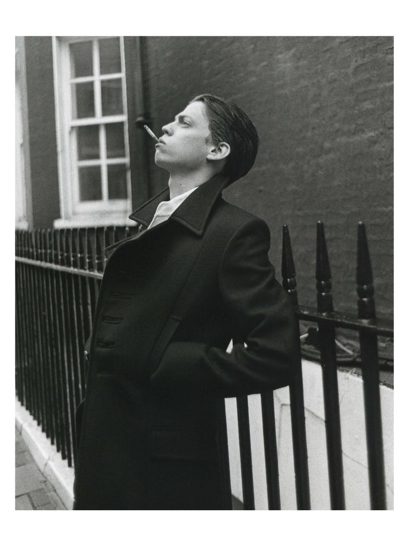 Young man in a dark double-breasted coat smoking against a black railing exterior