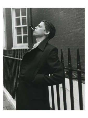 Young man in a dark double-breasted coat smoking against a black railing exterior