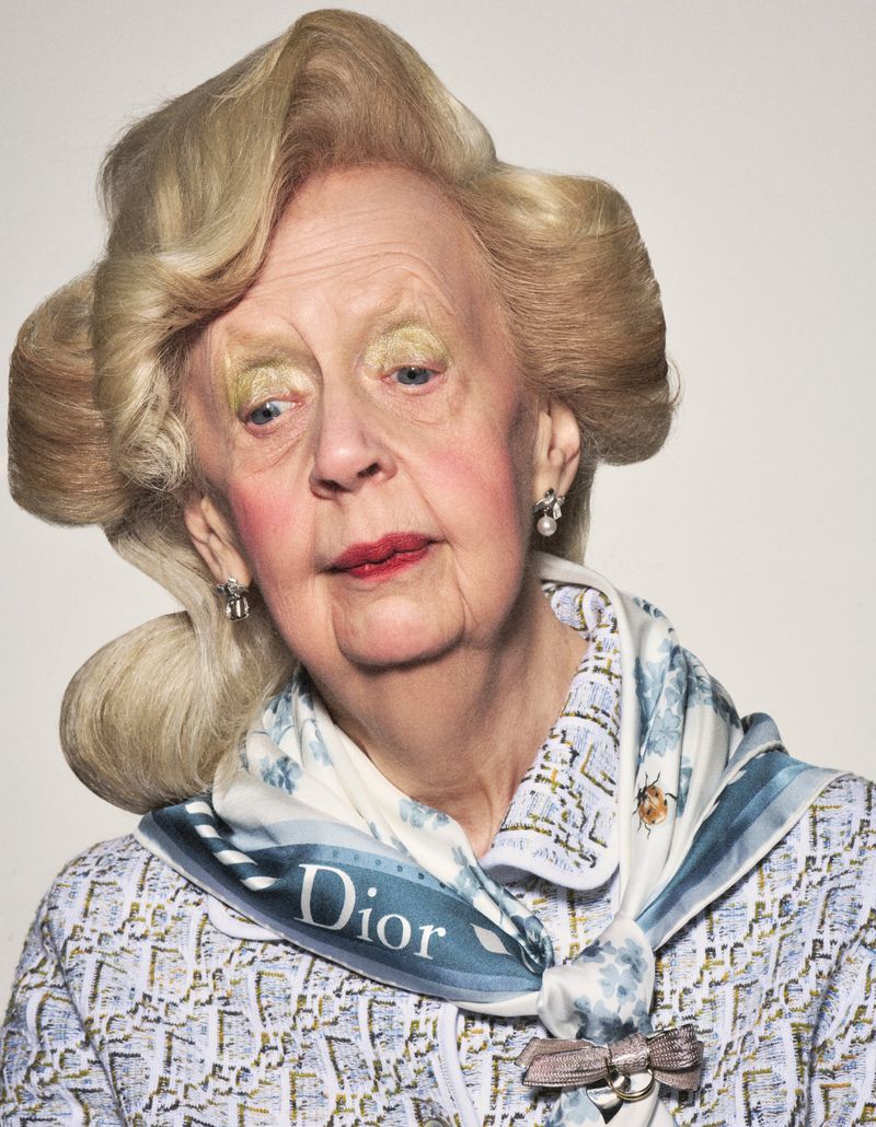 Close-up portrait of an elderly woman wearing a patterned Dior scarf and matching top with voluminous blonde hair and bold makeup.