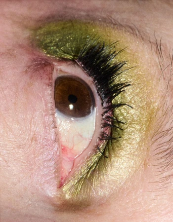 Extreme close-up of a brown eye featuring shimmering olive green eyeshadow, dark eyelashes, and precise eyeliner.