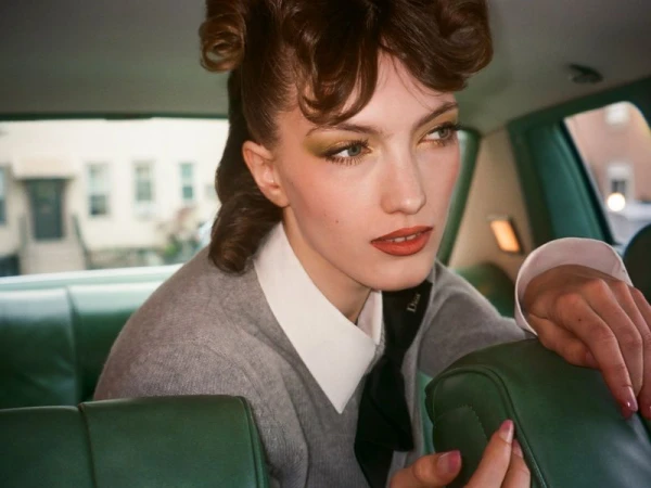 Model with vintage-styled hair seated inside a green car wearing a grey sweater and white collared shirt.