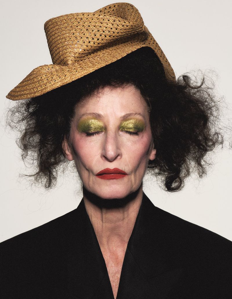 Close-up portrait of a woman wearing a tilted straw hat, green eyeshadow, red lipstick, and a black jacket against a plain background.