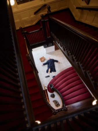 High angle shot of a man lying flat on a marble floor at the bottom of a grand staircase with red carpet and dark wooden railings.