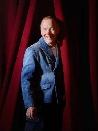 Portrait of David Sedaris wearing a blue denim jacket with a red clown nose, standing between red stage curtains.