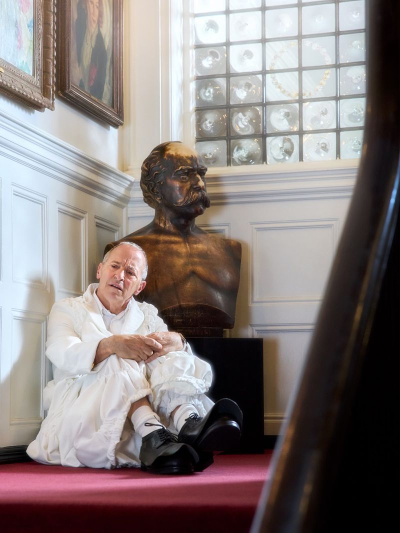 David Sedaris seated on red carpet in white garments next to a bronze bust for Zeit Magazine