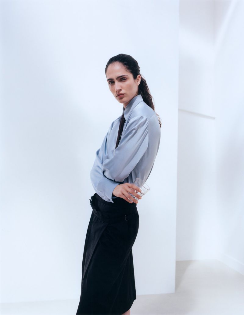 Model Sara Caballero in a light blue button-down shirt and black skirt against a white studio background for Muse Magazine.