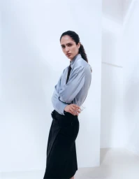 Model Sara Caballero in a light blue button-down shirt and black skirt against a white studio background for Muse Magazine.