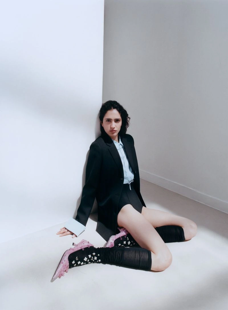 Model sitting on the floor against a white wall wearing a dark blazer, light blue shirt, shorts, knee-high socks, and pink heels