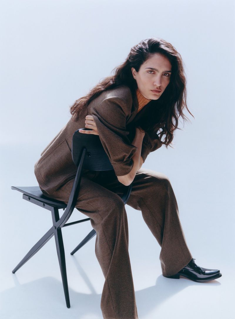 Model Sara Caballero seated backward on a black chair wearing a brown suit for Muse Magazine photographed by Cornelius Kaess.
