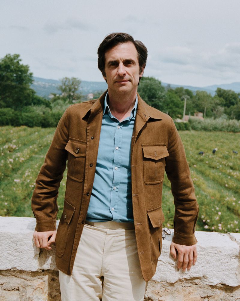 Man wearing a brown utility jacket and blue shirt leaning on a low white wall outdoors with a rural background.
