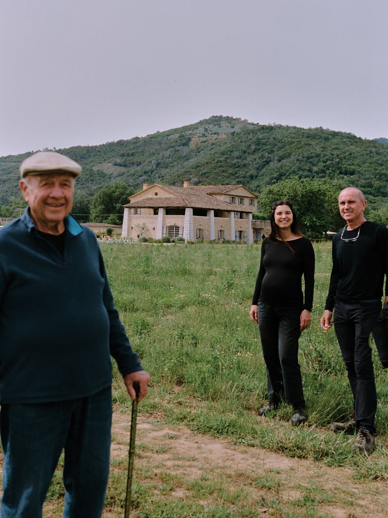 Elderly man in flat cap standing in foreground with walking stick, with two people and a large stone house in grassy field background.