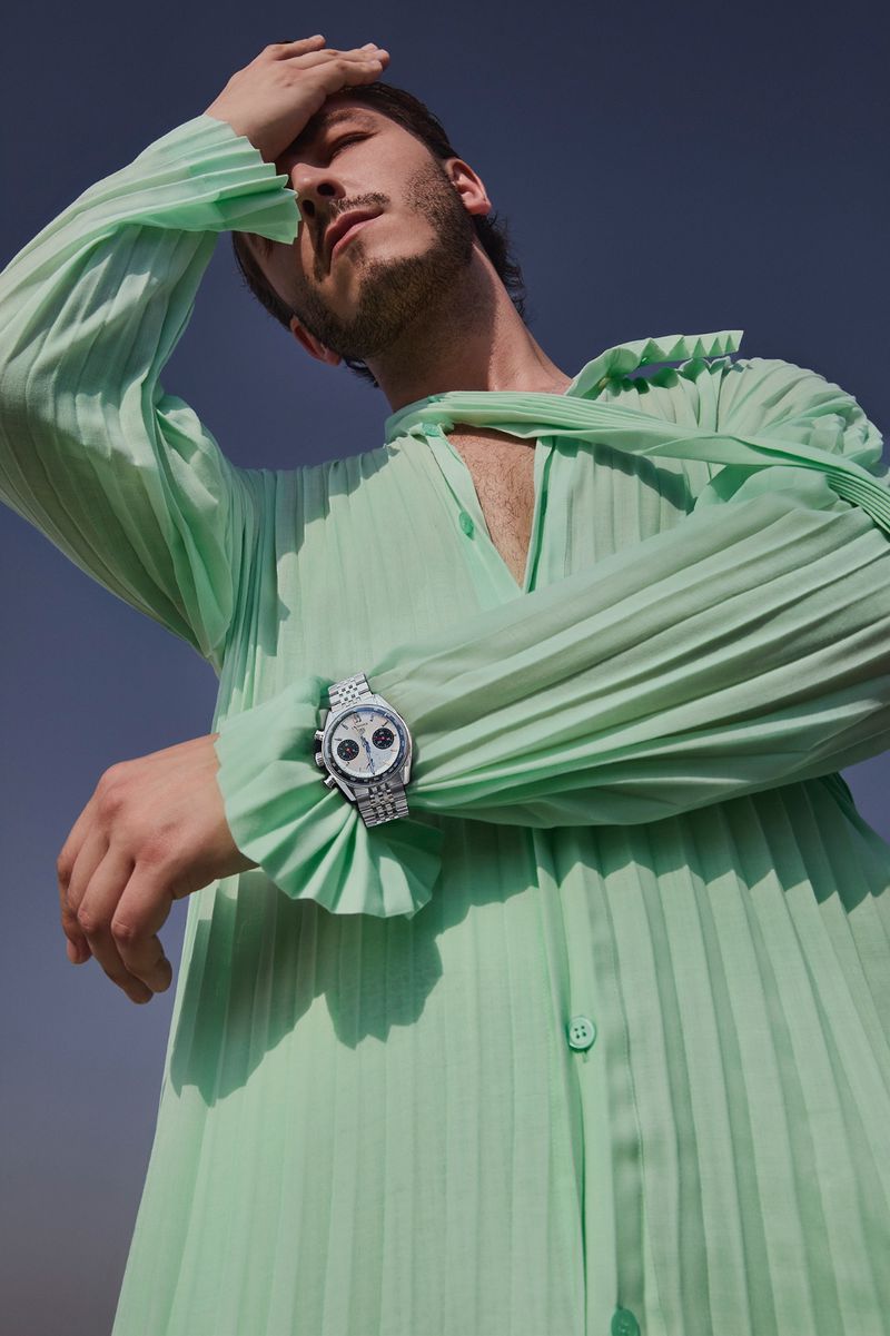Stylish man in a pleated mint green shirt poses in an outdoor setting with a watch on his wrist against a blue sky.