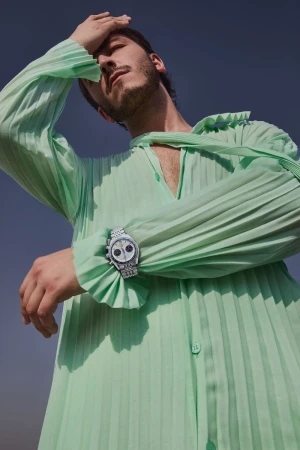 Stylish man in a pleated mint green shirt poses in an outdoor setting with a watch on his wrist against a blue sky.
