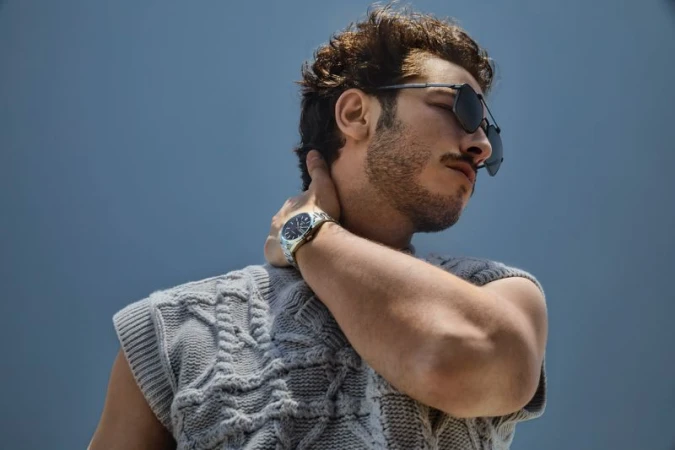 Portrait of a man in sunglasses wearing a grey cable knit sweater, touching his neck, posing against a blue gradient background.
