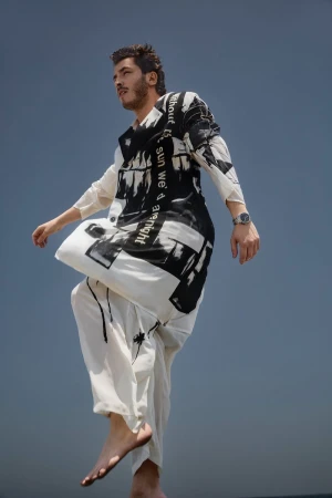 A man in a black-and-white patterned outfit is in motion against a muted blue sky. He's barefoot, creating a dynamic fashion statement.