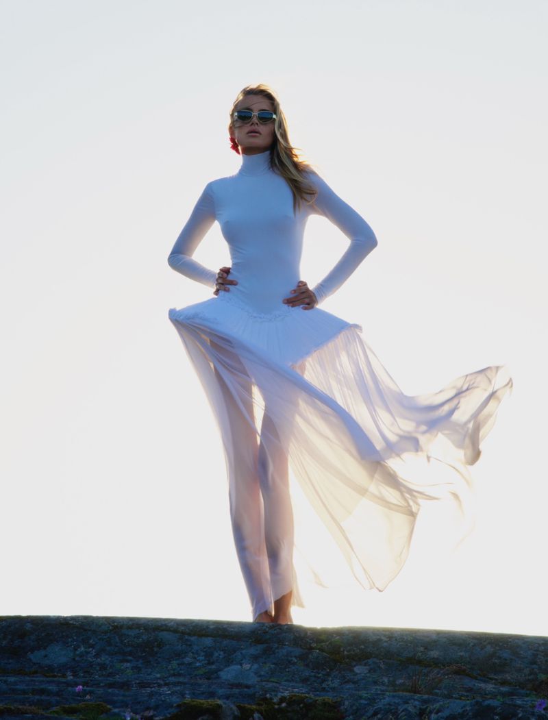 Model Emma Ellingsen wearing a white outfit and sunglasses against a bright sky background for Costume Norway.