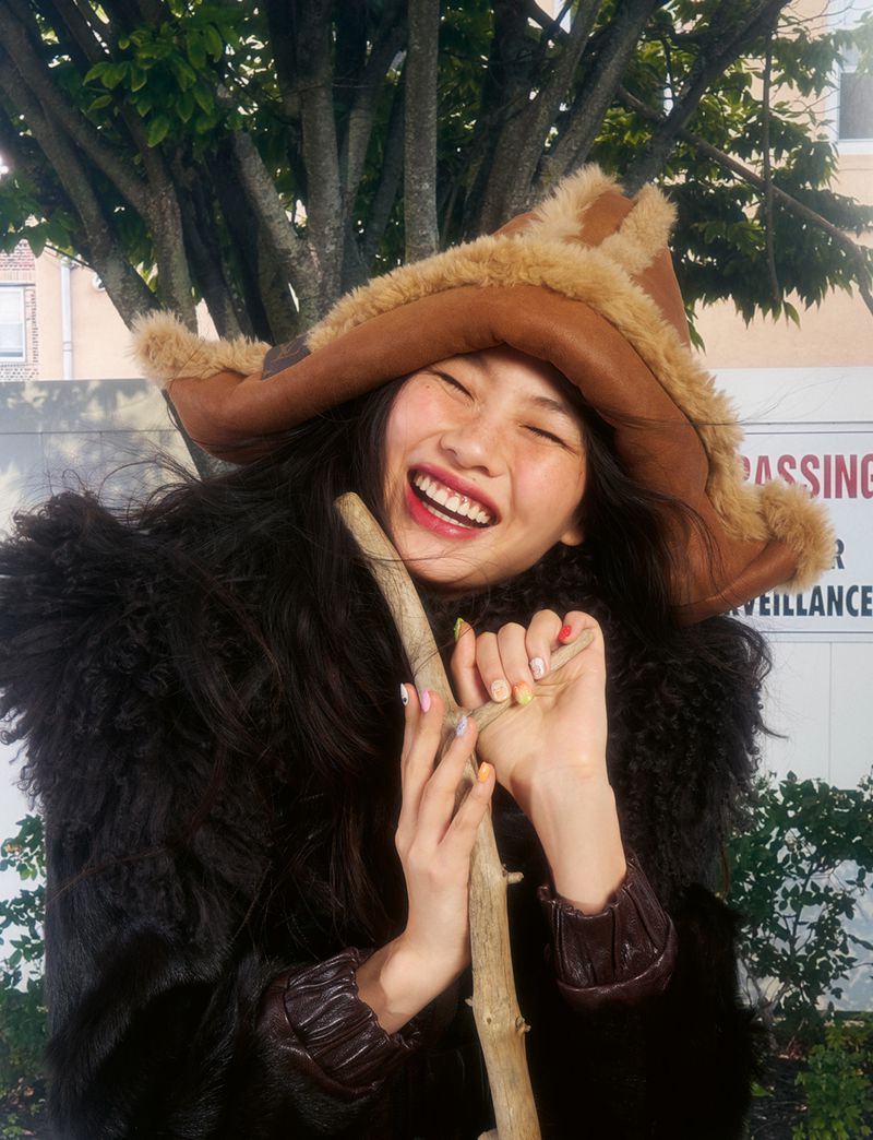 Hoyeon in a shearling hat and dark furry coat laughing while holding a wooden stick outdoors for AnOther Magazine.