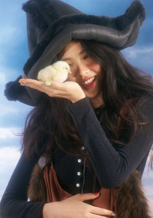 Smiling woman wearing a large dark hat holding a yellow chick in her hand against a blue sky backdrop.