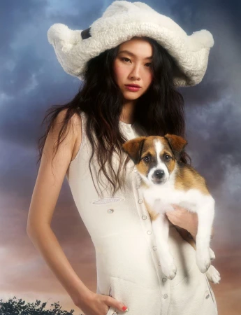 Asian woman holding a small puppy while wearing a white fluffy hat and a light-colored sleeveless dress against a cloudy sky backdrop.