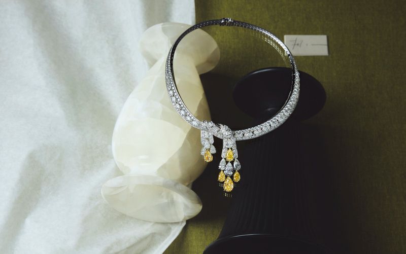Diamond necklace with yellow gemstones displayed on a green surface alongside a white stone vase and a black stand.