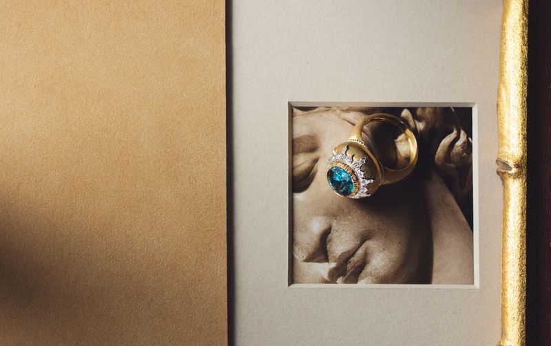 Close-up product shot of a gold ring with a blue gemstone resting on a photograph of a classical sculpture