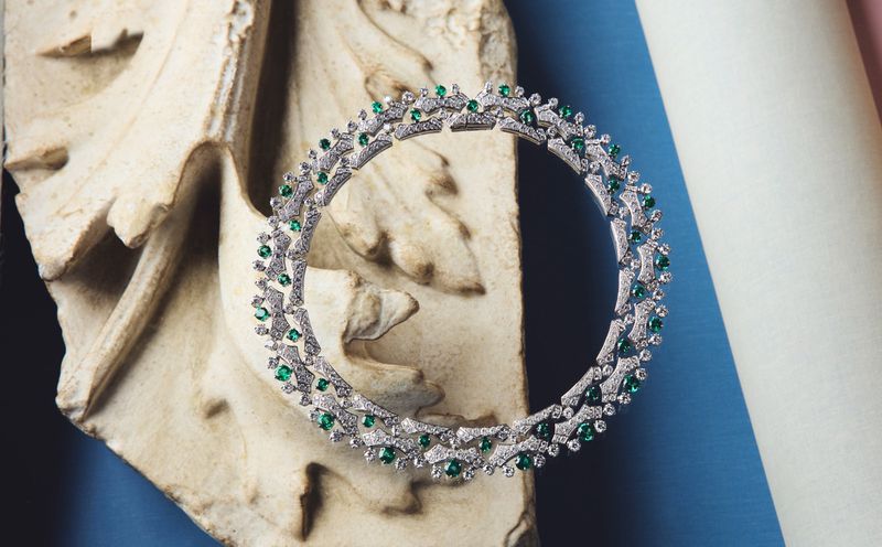 Close-up product shot of a diamond and emerald necklace displayed on an ornate stone architectural fragment against blue and pink pillars.