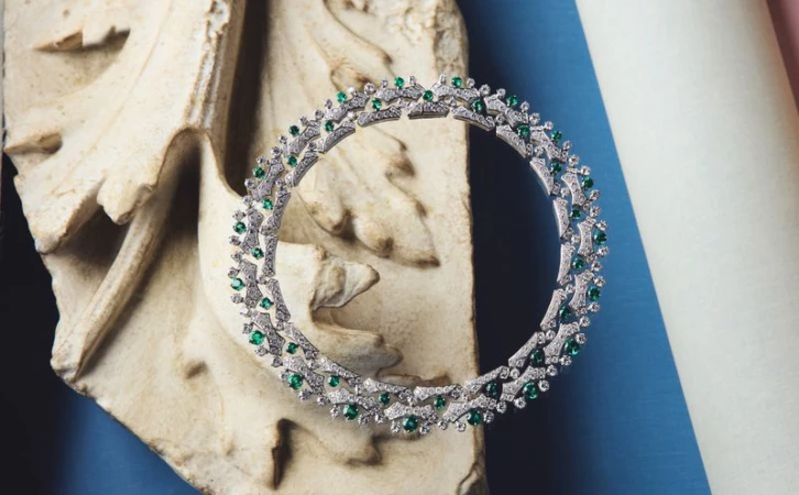 Close-up product shot of a diamond and emerald necklace displayed on an ornate stone architectural fragment against blue and pink pillars.